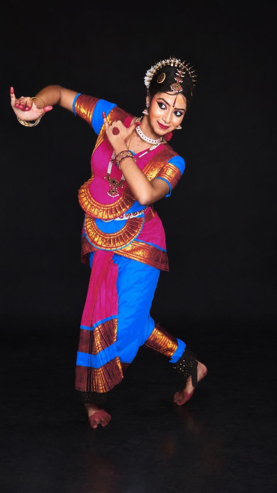 A Bharatanatyam dancer in costume, captured mid-pose against a black studio backdrop